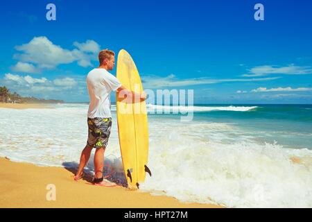 Homme avec une planche de surf sur la plage au coucher du soleil. Banque D'Images