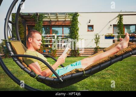 Jeune homme lisant un livre (apprentissage) à la balançoire dans le jardin de la maison familiale moderne. Banque D'Images