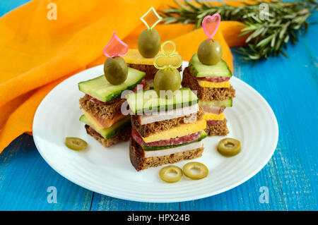 Mini-sandwiches (canape) avec des concombres frais, jambon, fromage, olives, salami, pain noir sur une plaque blanche sur fond bleu. Banque D'Images