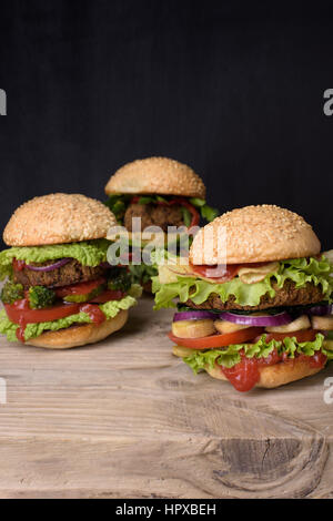 Hamburgers juteux avec de succulents petits pains de sésame sur les boulettes de boeuf et salade fraîche ingrédients sur une table en bois rustique, un arrière-plan sombre. Banque D'Images