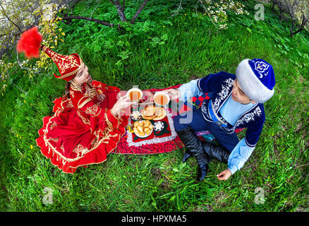 Couple en costume Kazakh assis dans le jardin en fleurs du printemps à Apple de pique-nique et de boire le thé à Almaty, Kazakhstan, en Asie centrale Banque D'Images