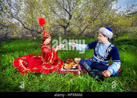 Couple en costume Kazakh assis dans le jardin en fleurs du printemps à Apple de pique-nique et de boire le thé à Almaty, Kazakhstan, en Asie centrale Banque D'Images