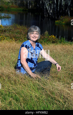 Femme se détend et profite de l'extérieur, dans le Nord de la Louisiane. Elle est à genoux dans l'herbe haute en face d'un étang. Banque D'Images