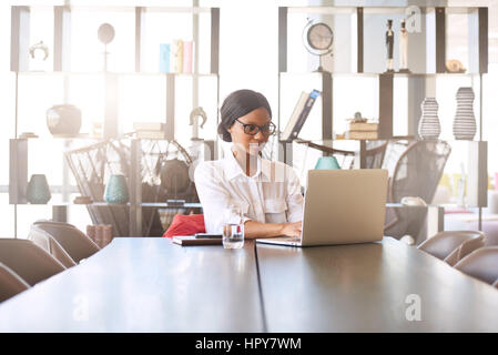 Belle jeune femme black bien instruits professionnels occupés à taper sur son ordinateur portable assis à sa table de salle à manger à la maison avec un grand écran Banque D'Images