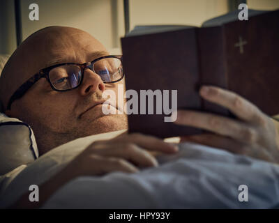 Middle aged man reading Bible on bed Banque D'Images