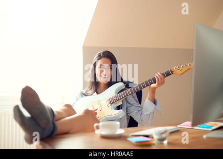 Jeune femme jouant de la guitare tout en gardant ses pieds sur la table avec la lumière du soleil dans la fenêtre derrière elle Banque D'Images