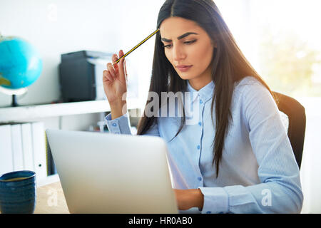 Young Asian businesswoman fronçant avec préoccupation qu'elle essaie de comprendre quelque chose qu'elle est en train de lire sur son ordinateur portable se gratter la tête avec il Banque D'Images