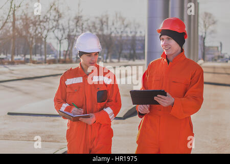 Cheerful young builders en orange uniforme avec la tablette et le bloc-notes. Tourné en extérieur horizontal. Banque D'Images