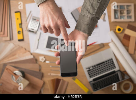 Décorateur accueil's hands holding a mobile phone à écran tactile, avec des outils de bureau, des ordinateurs portables et des échantillons de bois sur arrière-plan, Vue de dessus Banque D'Images