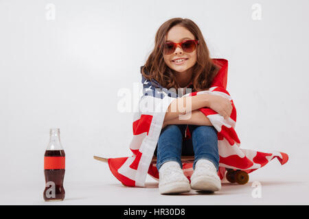 Profitant de la fête nationale. Jeune fille élégant sourire exprimant la joie tout en étant assis sur la planche à roulettes à l'arrière-plan blanc et souriant Banque D'Images