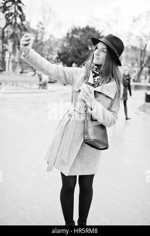 Modèle jeune fille dans un manteau gris et noir chapeau avec sac à main en cuir sur les épaules restent avec tasse de café en plastique et faire en selfies rue de ville. B Banque D'Images