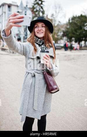 Modèle jeune fille dans un manteau gris et noir chapeau avec sac à main en cuir sur les épaules restent avec tasse de café en plastique et faire en selfies rue de ville. Banque D'Images