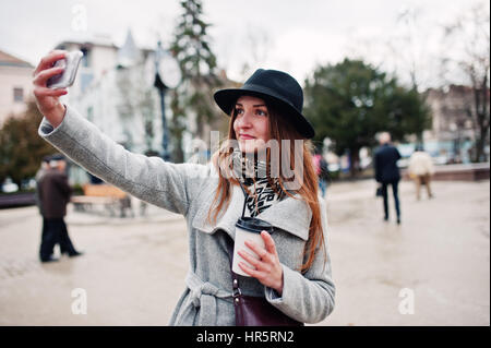 Modèle jeune fille dans un manteau gris et noir chapeau avec sac à main en cuir sur les épaules restent avec tasse de café en plastique et faire en selfies rue de ville. Banque D'Images