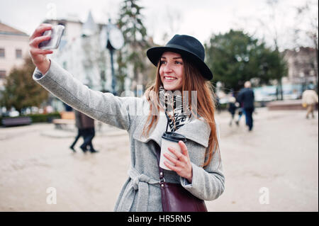 Modèle jeune fille dans un manteau gris et noir chapeau avec sac à main en cuir sur les épaules restent avec tasse de café en plastique et faire en selfies rue de ville. Banque D'Images