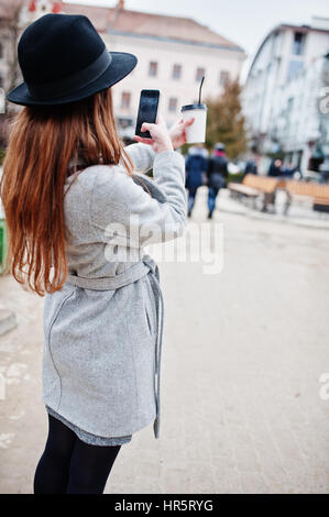 Modèle jeune fille dans un manteau gris et noir chapeau avec sac à main en cuir sur les épaules restent avec tasse de café en plastique et faire en selfies rue de ville. Banque D'Images