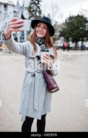 Modèle jeune fille dans un manteau gris et noir chapeau avec sac à main en cuir sur les épaules restent avec tasse de café en plastique et faire en selfies rue de ville. Banque D'Images