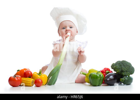 Petite fille vêtue de blanc et un tablier chef hat mange oignon frais. Enfant avec des légumes : aubergine, pop-corn, broccolli, poivrons, courgettes, tomates Banque D'Images