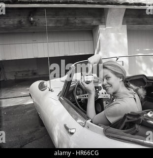 Années 1960, historique, dame dans le siège du conducteur d'une voiture de sport jaguar haut montre comment ouvrir les portes automatiques de son garage à l'aide d'un bouton new dispositif de commande à distance. Banque D'Images