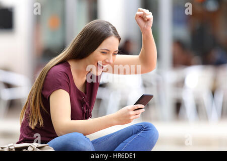 Femme étonné de recevoir de bonnes nouvelles en ligne maintenant un téléphone intelligent assis sur un banc dans la rue avec un milieu urbain Banque D'Images