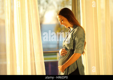 Femme enceinte en regardant son ventre près d'une fenêtre avec un accueil chaleureux à la maison de la lumière au coucher du soleil Banque D'Images