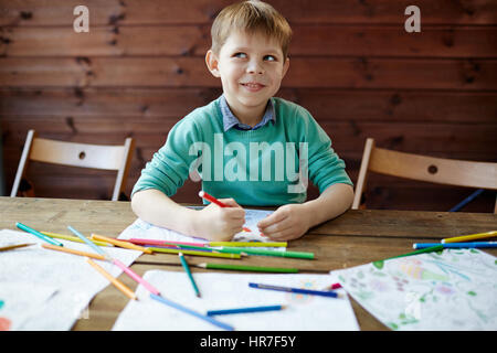 Cute little boy dans l'aigue-marine pour homme assis au bureau en bois avec des livres à colorier, des crayons et des feutres et regardant ailleurs rêveur Banque D'Images