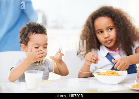 Mignon bébé garçon et sa jolie soeur avec de longs cheveux bouclés assis à table de dîner et manger de délicieux pâtes aux mains Banque D'Images