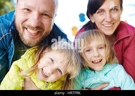 Head and shoulders portrait of happy family in park : les parents d'âge moyen dans les vêtements de sport à la caméra avec un sourire à pleines dents et s'étreindre leur drôle Banque D'Images