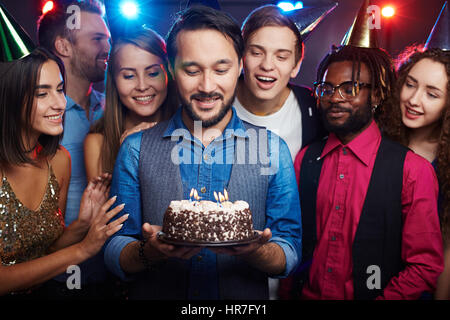 Jeune homme barbu debout parmi des amis proches, la tenue du gâteau d'anniversaire avec des bougies et faire un vœu Banque D'Images