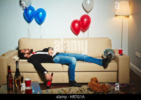 Smiling middle-aged man lying on couch et boire de la bière à partir de la bouteille dans un salon en désordre, bouteilles d'alcool vides et des gobelets en plastique partout permanent Banque D'Images