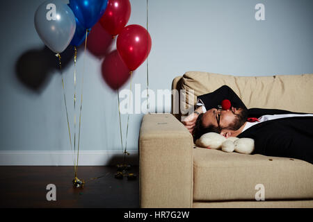 Portrait en taille de l'homme barbu avec nez de clown rouge couchage sur canapé après partie sauvage, l'ours blanc couché sous sa tête Banque D'Images