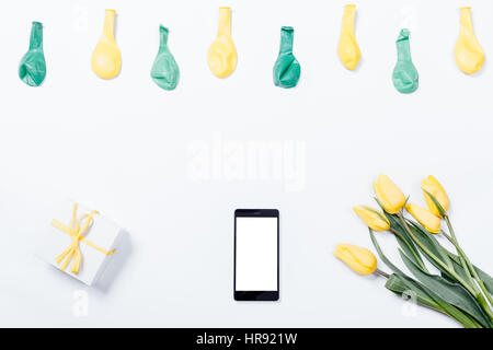 Fleurs jaunes, d'un téléphone mobile, petite boîte-cadeau avec un ruban et des ballons sur un tableau blanc, vue du dessus Banque D'Images