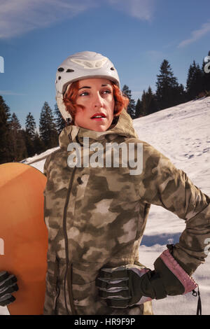 Par beau temps jusqu'à la taille d'une femme aux cheveux rouge avec casque, dans la pente, avec snowboard à part Banque D'Images