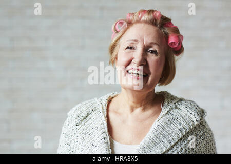 Joyful woman with hair curlers debout contre un mur en brique blanche et à l'écart avec sourire à pleines dents Banque D'Images