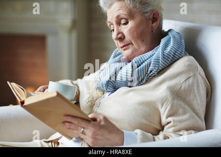 Calme soirée d'hiver par cheminée : Pretty woman s'asseoir sur un fauteuil avec livre intéressant et tenant en main une tasse de thé Banque D'Images