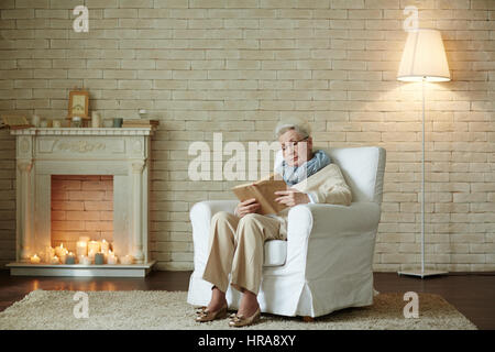 Portrait en pied d'une femme âgée à lunettes est concentré sur la sortie de lecture livre tout en restant assis dans un fauteuil confortable contre mur en brique blanche Banque D'Images
