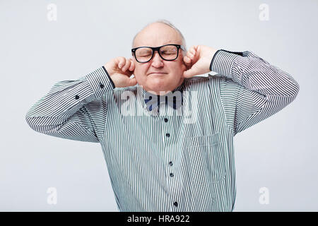 Jusqu'à la taille de l'élégant portrait homme âgé en chemise rayée avec papillon boucher ses oreilles avec les doigts et pinçant ses yeux Banque D'Images