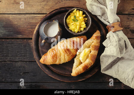 Le petit-déjeuner avec deux croissant, beurre, crème et de tranches de mangues, servi sur le bois servant à bord Rond de serviette textile sur fond de bois ancien. Banque D'Images
