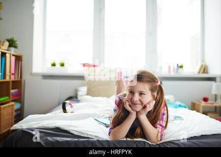 Jolie fille aux cheveux long avec queues de lying on bed with se pencha la tête dans ses mains, à la recherche sur le côté et souriant malicieusement Banque D'Images