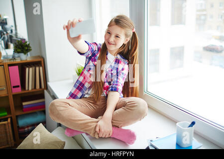 Jolie fille de l'école roux avec queues assis sur window-sill dans sa chambre à coucher et faire sur smartphone selfies Banque D'Images