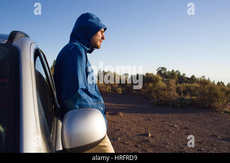 Jeune homme en veste bleue et le capot se reposer dans sa voiture, à la recherche sur le côté et en plissant les yeux en plein soleil Banque D'Images