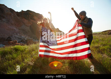 Couple de jeunes gens à sauter et à crier haut et fort la main, répandant big USA flag on top of hill dans les montagnes Banque D'Images