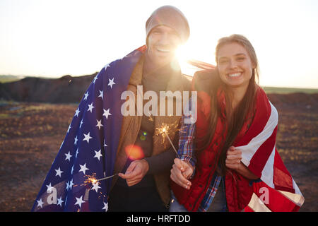 Portrait of young couple wearing fashionable vêtements touristiques rire et holding sparklers, aussi bien enveloppé dans le drapeau américain contre le soleil de mountai Banque D'Images