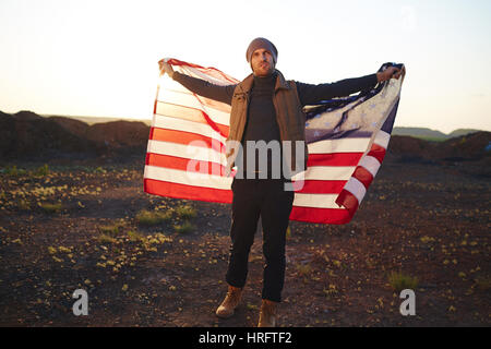 Portrait de beau jeune homme vêtu à la mode des vêtements de randonnée en montagne seul armes propagation holding big American bannière contre sunse Banque D'Images