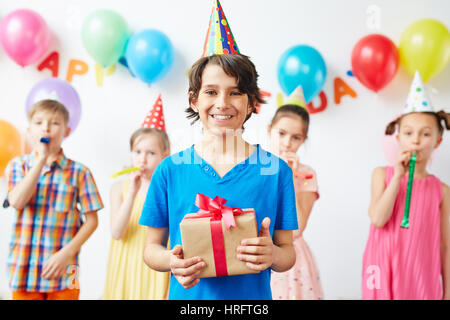 Portrait de heureux anniversaire boy holding fort à présent debout devant souriant joyeusement avec ses amis, les bruiteurs parti soufflant derrière dans backg Banque D'Images