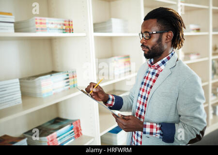 Voir le profil de l'homme barbu concentré avec de courtes dreadlocks debout à étagères et prise de notes avec crayon Banque D'Images