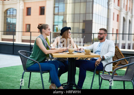 Groupe de jeunes gens d'affaires créatifs, deux jolies femmes et un homme, assis à une table pour discuter travail et souriant pendant pause café Banque D'Images