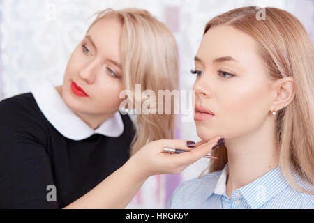 Cosmetologist blonde en robe noire et rouge à lèvres à la suite de procédure de miroir de maquillage permanent. Artiste de toucher des femmes face Banque D'Images