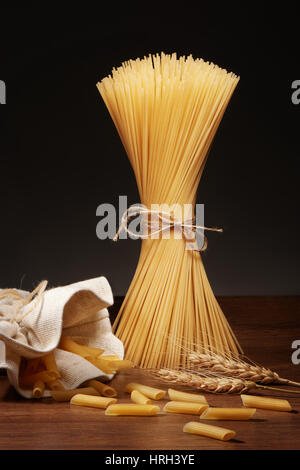 Les pâtes spaghetti sec attaché avec une corde, pâtes penne rigate tombe de son sac de toile et les épis de blé sur la table en bois sombre sur fond gris Banque D'Images