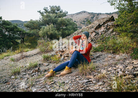 Portrait d'une belle jeune femme blonde avec chapeau, pull rouge, bleu jeans et bottes jaunes se trouve en plein air, sur l'arrière-plan de montagnes et des forêts Banque D'Images