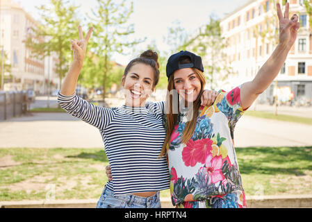 Deux jeunes étudiantes heureux faire v-signes tels qu'ils sont dans les bras sur une paisible rue urbaine sur une chaude journée d'été smiling at the camera Banque D'Images